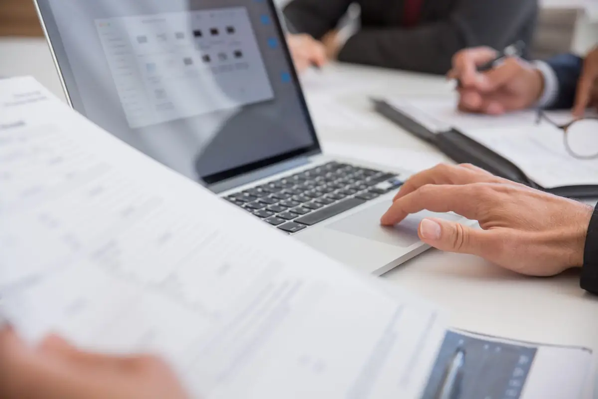 Hands reviewing printed financial documents beside a laptop.